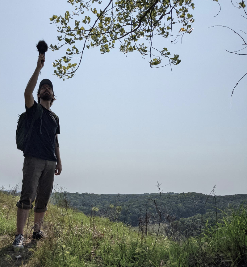 a photo of me standing on a hill, my arm stretched up high above my head, holding a small field recorder. It looks like I'm trying to record the sounds of a tree branch. Behind me is a clear sky, on a hot day, over some tree-covered hills.